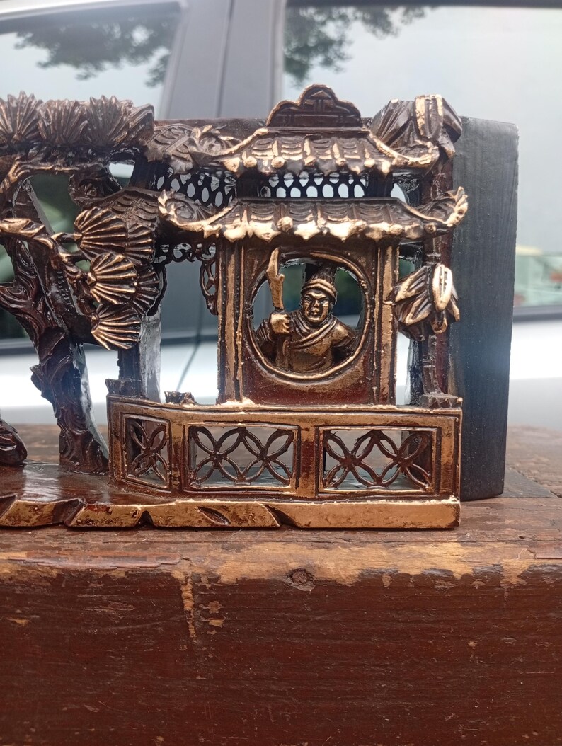 May include: A dark brown and gold-toned decorative carving featuring a detailed architectural structure with a tiled roof and a circular window. Inside the window, a figure holds a polearm. To the left, a tree with fan-shaped leaves is depicted.