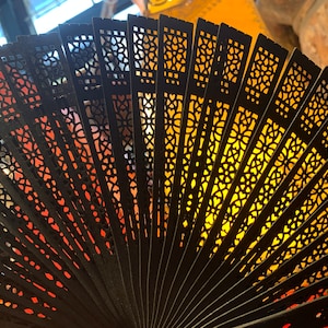 May include: A black folding fan with intricate cut-out designs. The fan is open and the light shines through the cut-outs, creating a pattern of light and shadow.