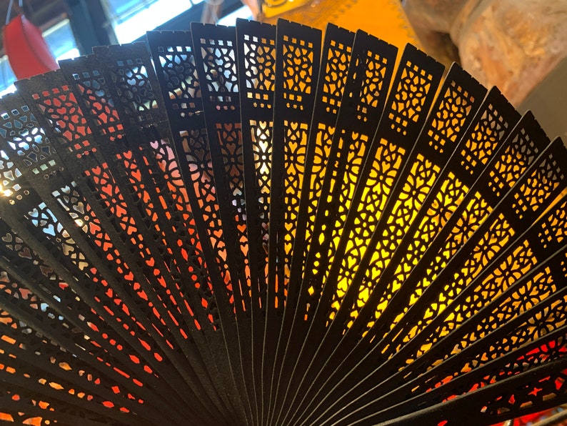 May include: A black folding fan with intricate cut-out designs. The fan is open and the light shines through the cut-outs, creating a pattern of light and shadow.