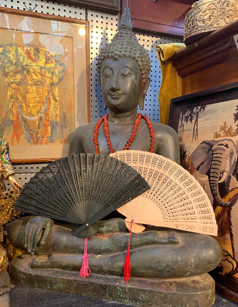 May include: A bronze Buddha statue with a red beaded necklace sits with two hand fans, one black and one white, resting on its lap. The statue is in a shop setting with other items in the background.