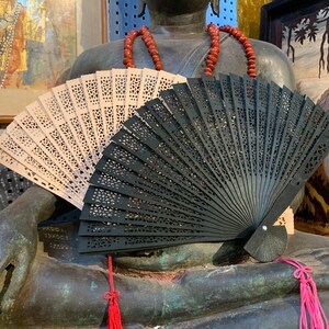 May include: Two hand fans, one black and one beige, are resting on the lap of a large bronze statue. The fans are made of wood and have intricate designs.