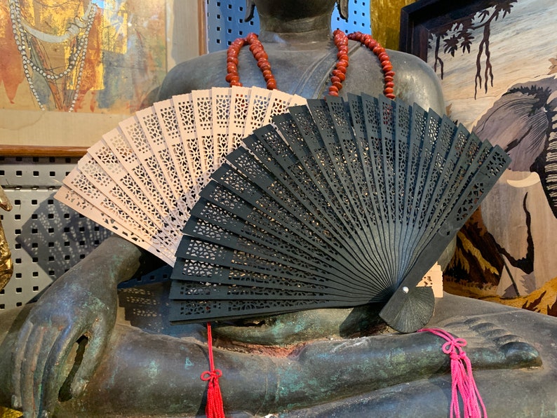 May include: Two hand fans, one black and one beige, are resting on the lap of a large bronze statue. The fans are made of wood and have intricate designs.