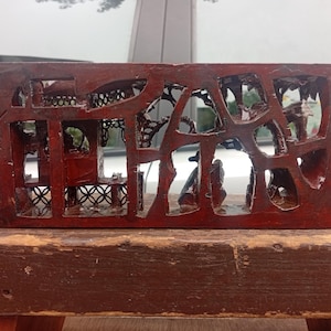 May include: A rectangular, dark reddish-brown wooden block featuring intricate, openwork carvings of architectural structures and foliage. The surface has a glossy finish, and the left side displays etched numerical markings, specifically 101 and 522.