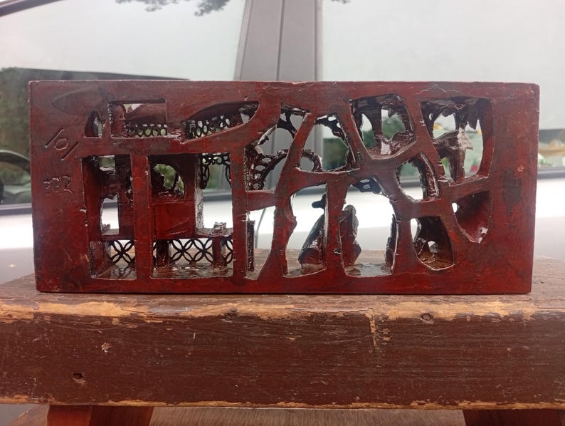 May include: A rectangular, dark reddish-brown wooden block featuring intricate, openwork carvings of architectural structures and foliage. The surface has a glossy finish, and the left side displays etched numerical markings, specifically 101 and 522.