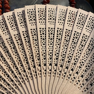 May include: A white folding fan with intricate cut-out designs. The fan is made of thin, delicate wood and has a traditional Asian style.