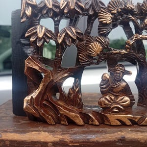May include: A dark brown and gold-toned carved wooden relief panel featuring a seated figure in traditional attire positioned beneath stylised trees and rock formations. The intricate openwork design highlights the contrast between the dark wood and the gold-painted raised details on the figure and foliage.