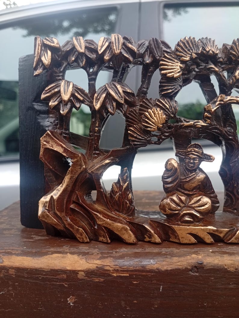 May include: A dark brown and gold-toned carved wooden relief panel featuring a seated figure in traditional attire positioned beneath stylised trees and rock formations. The intricate openwork design highlights the contrast between the dark wood and the gold-painted raised details on the figure and foliage.