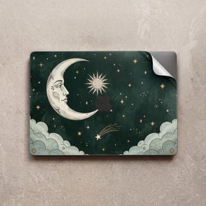 May include: A dark green laptop skin featuring a crescent moon with a human face, a stylized sun, a shooting star, and various small stars. The design includes light blue clouds at the bottom corners and a cutout for an Apple logo.