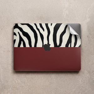 May include: A laptop skin featuring a zebra print design in black and cream, with a solid burgundy bottom half. The Apple logo is visible. The top right corner of the skin is peeling back.