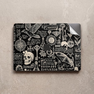 Könnte beinhalten: Schwarze Laptop-Hülle mit einem detaillierten, gotisch inspirierten Design. Die Kunstwerke umfassen einen Raben, einen Schädel, Schlüssel, einen Kompass und verschiedene alchemistische Symbole. Das Wort "MAGNUS" wird in einem Banner angezeigt, und "SAPIENTIA" steht unter einem Schädel.