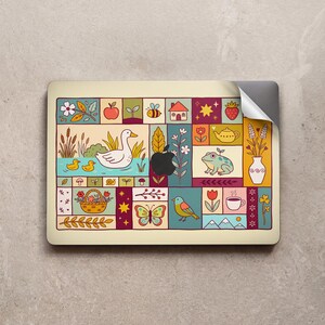 Puede incluir: Una pegatina rectangular para portátil con una cuadrícula de ilustraciones coloridas que incluyen un pato blanco, dos patitos, una rana, una mariposa, un pájaro, una tetera, una taza de café, una fresa, una manzana, una casa, flores y plantas sobre un fondo crema.