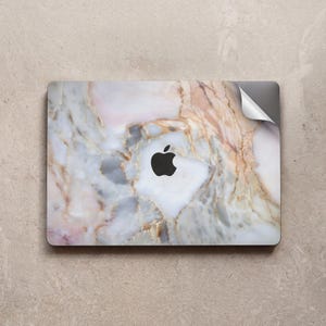 May include: A laptop skin featuring a marble design with white, pink, and brown veining. The Apple logo is visible in black. The skin is applied to a laptop, with a corner of the skin peeling up.