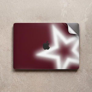 May include: A rectangular laptop skin featuring a deep maroon background with a large, soft-focus white star graphic. A black Apple logo is centered on the surface, and the top right corner of the adhesive skin is partially peeled back.