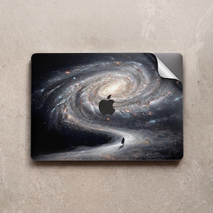 Op de afbeelding: Laptop skin met een wervelend melkwegstelsel ontwerp en een silhouet dat over een pad loopt. De skin heeft een zwart Apple logo en een gedeeltelijk losgemaakte hoek.