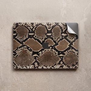 May include: A laptop skin featuring a brown and black snakeskin pattern with a realistic scale texture. The rectangular adhesive cover is partially peeled back at the top right corner, revealing the metallic surface of a laptop underneath.