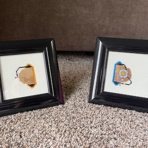 May include: Two framed cross-stitch artworks. Each features a geometric design in tan, orange, and black on a white background. The frames are square and black. The artworks are displayed on a neutral-colored carpet.