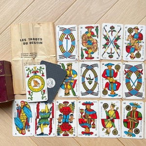 May include: A vintage tarot card set, featuring a burgundy box labeled "Arcanes du Destin" and a paper sleeve with French text. The cards display colorful illustrations and symbols, with a focus on the "Les Tarots du Destin" theme.