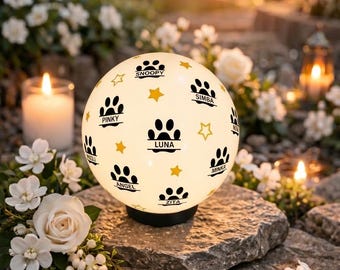 May include: A glowing, spherical outdoor light with paw prints and star designs. The names "Snoopy", "Luna", "Simba", "Pinky", "Angel", "Minka", and "Zita" are printed on the light. The light is set on a stone surface, surrounded by white flowers and candles.