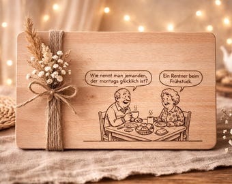 May include: Wooden cutting board with a humorous engraved illustration of an elderly couple at breakfast, with text bubbles. The board is decorated with a twine-wrapped dried flower arrangement. The background is blurred with warm lights.