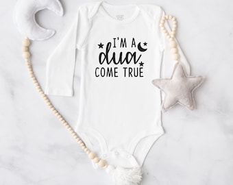 May include: White baby onesie with long sleeves, featuring the text "I'M A dua COME TRUE" in black. The onesie is surrounded by a wooden bead garland, a plush star, and a crescent moon. A cute baby gift.