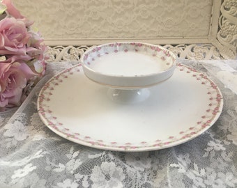 May include: A white ceramic cake stand with a floral design. The stand has a round base and a smaller round top with a pedestal in the middle. The floral design is pink and green.