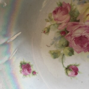K&ouml;nnte beinhalten: Eine antike Porzellanschale mit einem zarten Blumendesign mit rosa Rosen und gr&uuml;nen Bl&auml;ttern auf hellblauem Hintergrund. Die Schale hat eine irisierende Glasur, die einen Regenbogeneffekt erzeugt.