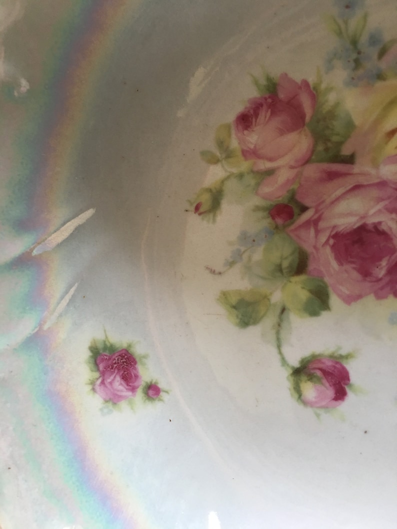 K&ouml;nnte beinhalten: Eine antike Porzellanschale mit einem zarten Blumendesign mit rosa Rosen und gr&uuml;nen Bl&auml;ttern auf hellblauem Hintergrund. Die Schale hat eine irisierende Glasur, die einen Regenbogeneffekt erzeugt.