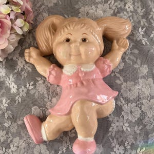 May include: A ceramic doll figurine in a playful pose, featuring a light skin tone, rosy cheeks, and two pigtails. The doll wears a pink dress with a white collar and matching pink shoes. The figurine is designed to be mounted on a wall.