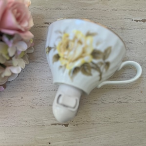 May include: A white porcelain teacup with a gold rim and a floral design featuring yellow roses. The cup has a handle and a small base.