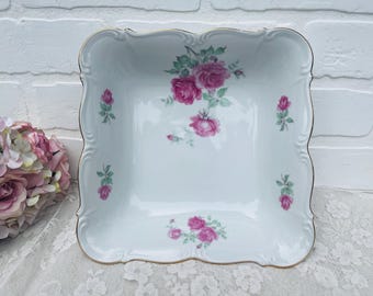 May include: A white square serving dish with a scalloped edge and gold trim. The dish is decorated with pink roses and green leaves. A floral arrangement is visible in the background.