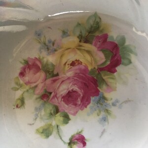 K&ouml;nnte beinhalten: Eine wei&szlig;e Porzellanschale mit einem Blumendesign aus rosa und gelben Rosen und blauen Vergissmeinnicht. Die Schale hat einen gewellten Rand und eine gl&auml;nzende Oberfl&auml;che.