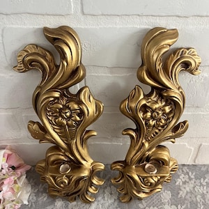 May include: A pair of gold-tone metal wall sconces with ornate floral designs. Each sconce has a single candle holder.