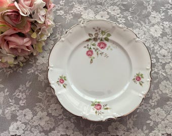 May include: A white porcelain plate with a delicate floral design featuring pink roses and green leaves. The plate has a scalloped edge and a gold rim.