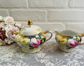 May include: A vintage porcelain sugar bowl and creamer set with a floral design. The set features a white background with hand-painted pink, yellow, and green roses. The sugar bowl has a gold lid and a gold trim around the rim. The creamer has a gold handle and a gold trim around the rim.