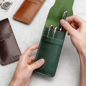 Pu&ograve; includere: Una collezione di astucci in pelle per penne in vari colori, tra cui marrone e verde. Un astuccio &egrave; aperto, rivelando penne all'interno. Gli astucci sono personalizzati con iniziali. Sono visibili anche una bussola e una penna.