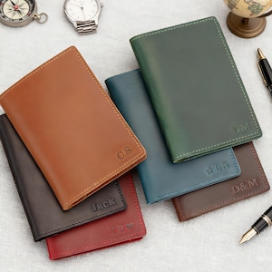 Könnte beinhalten: Eine Auswahl an personalisierten Leder-Reisepasshüllen in verschiedenen Farben, darunter Braun, Grün und Rot. Jede Hülle ist mit Initialen oder Namen versehen, im Hintergrund sind Kompass, Uhr und Globus zu sehen.