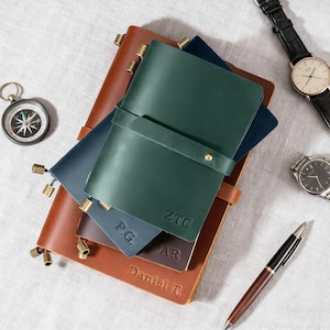 May include: A selection of leather journals in brown, green, and blue, with gold-tone metal accents. A compass, two watches, and a pen are also present. The journals are embossed with initials and names, a stylish stationery set.