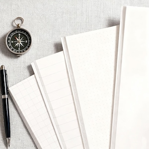 Op de afbeelding: Een zilveren kompas, zwarte pen en verschillende witte notitieboeken met verschillende paginastijlen zijn gerangschikt op een getextureerd, lichtgrijs oppervlak. De notitieboeken hebben gelinieerde, geruite en gestippelde pagina's. Het kompas is rond met een zwarte en groene wijzerplaat.