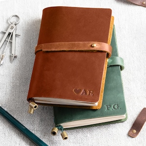 May include: Two leather journals stacked together. The top journal is brown with a horizontal strap, a brass stud, and the embossed text AR with a heart symbol. The bottom journal is green with the embossed text P.G. Both have brass charms.