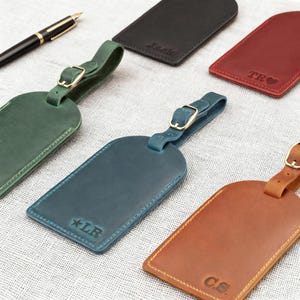 Könnte beinhalten: Eine Auswahl an personalisierten Leder-Gepäckanhängern in verschiedenen Farben, darunter Grün, Blau und Braun. Jeder Anhänger hat einen Schnallenriemen und ist mit Initialen oder einem Namen geprägt. Ein schwarzer Stift ist ebenfalls zu sehen.