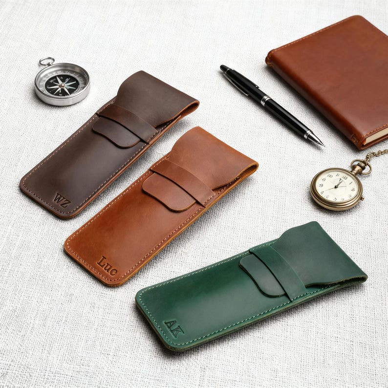 Pu&ograve; includere: Tre astucci in pelle per penne marroni, beige e verdi, ciascuno con chiusura a patta e iniziali personalizzate. Sono visibili anche una bussola, un orologio da tasca, una penna e un taccuino in pelle.