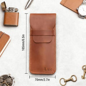 Pu&ograve; includere: Un astuccio per orologio in pelle marrone, alto 165 mm e largo 70 mm, con il nome "Luc" impresso sul davanti. L'astuccio ha una chiusura a patta e un cinturino. Altri accessori includono un accendino, una penna, un orologio e delle chiavi.