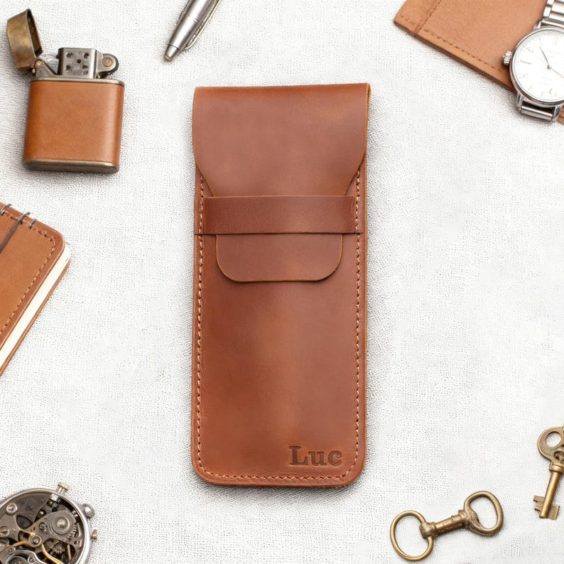 Pu&ograve; includere: Una custodia per telefono in pelle marrone con chiusura a patta e bordi cuciti. La parola "Luc" &egrave; impressa in basso. Altri oggetti includono un accendino, una penna, un orologio e chiavi.