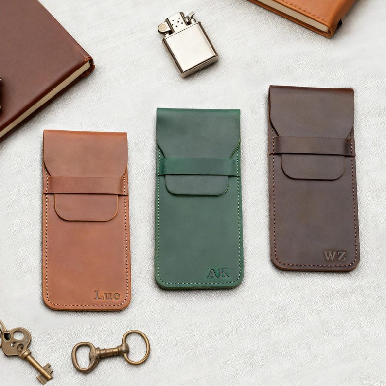 Pu&ograve; includere: Tre custodie in pelle per telefono, marrone, verde e marrone scuro. Ogni custodia ha una chiusura a patta e un bordo cucito. La custodia marrone &egrave; impressa con "Luc", quella verde con "AK" e quella marrone scuro con "WZ". Sono visibili anche un accendino argentato e un taccuino marrone.