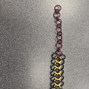 May include: A chainmaille strip featuring a wide section of interlocking black and yellow rings that transitions into a narrower tail of alternating red and black rings. The piece is constructed from circular metal links in a repeating pattern.