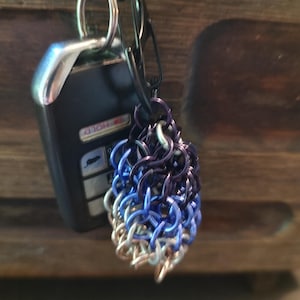May include: A handmade keychain featuring a chainmail design in shades of silver, blue, and purple. The keychain is attached to a black carabiner clip and a car key. The chainmail is a decorative accessory.