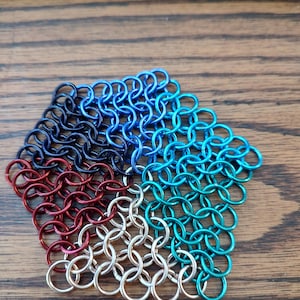 May include: A pentagon-shaped chainmail piece composed of interlocking metal rings. The chainmail is divided into five sections, each with a different color: black, blue, teal, silver, and red. The piece is displayed on a wooden surface.