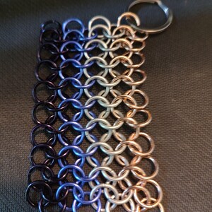 May include: A rectangular chainmail keychain with a gradient of colors. The left side is dark purple, transitioning to blue, silver, and then copper on the right. A silver key ring is attached to the top right corner.