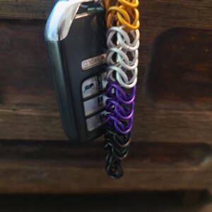 May include: A car key fob with a silver key ring and a chainmail keychain. The keychain is made of interlocking metal rings in a gradient of colors: gold, white, purple, and black. The key fob is black with silver accents.