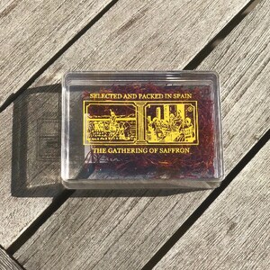 May include: A clear plastic box containing red saffron threads. The box has a gold label with the text "SELECTED AND PACKED IN SPAIN" and "THE GATHERING OF SAFFRON".